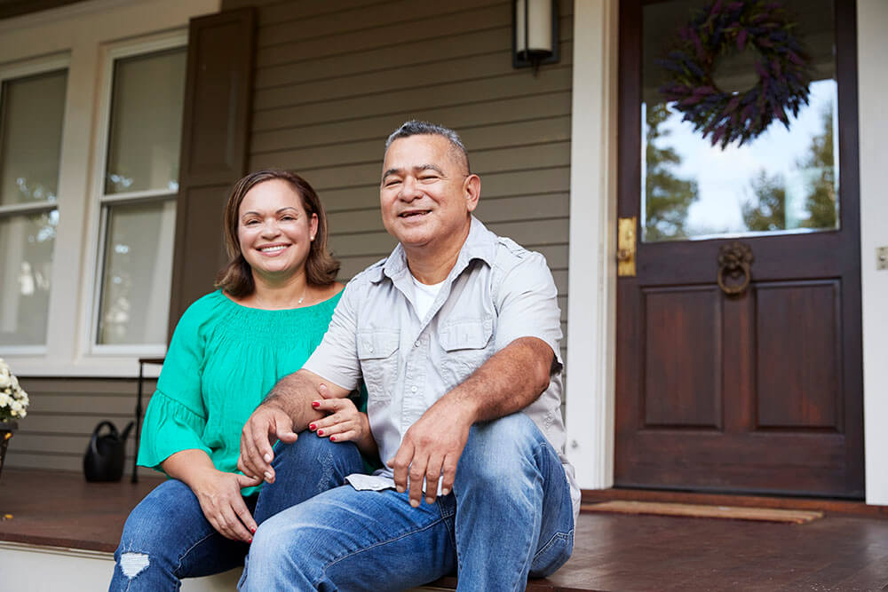 Portrait Of Smiling Senior Couple Sitting In Front Of Their Home.